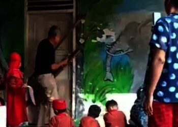 PENYEGELAN–Seorang lelaki paruh baya, sedang memasang dua bilah kayu, dengan paku pada kusen pintu masuk ruang kelas. Sementara itu, anak – anak usia dini yang mengenakan seragam terlihat ada yang berdiri dan duduk diteras sekolah, menyaksikan aksi penyegelan tersebut, Minggu (26/9/2021). (ISTIMEWA)