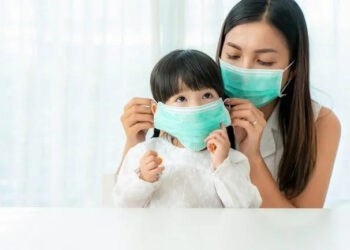 Asian mother wearing wearing to her daughter healthy face mask sitting in living room at home to prevent PM2.5 dust, smog, air pollution and COVID-19. Healthcare concept.